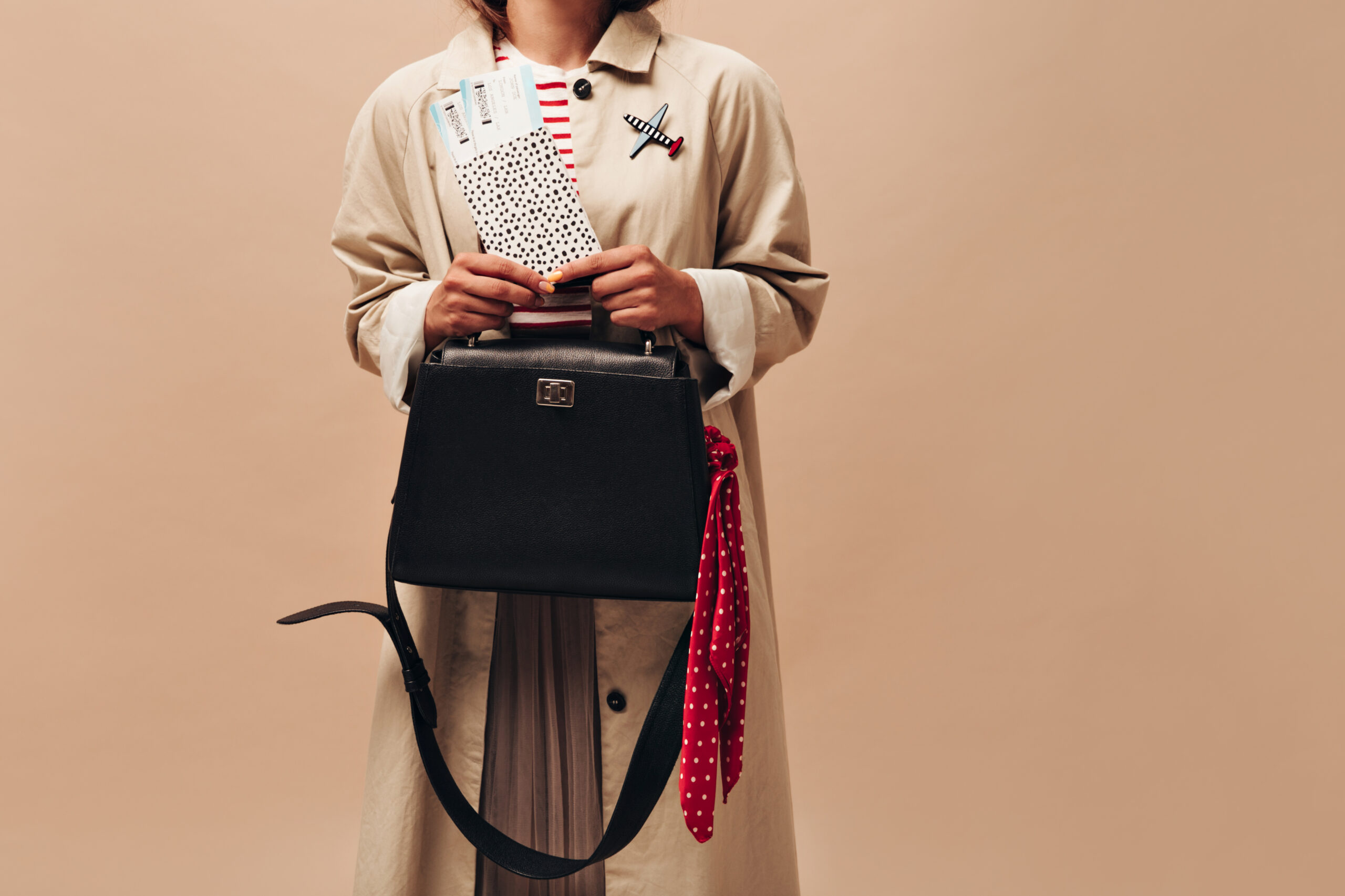 Stylish woman in long autumn coat and modern striped sweater holds black handbag and tickets on isolated beige background..